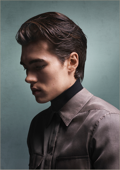 Young man with a modern precision haircut and style, featuring slicked-back brown hair, wearing a button-up shirt and black turtleneck, looking down against a muted teal background. - Garbo A Salon & Spa TX