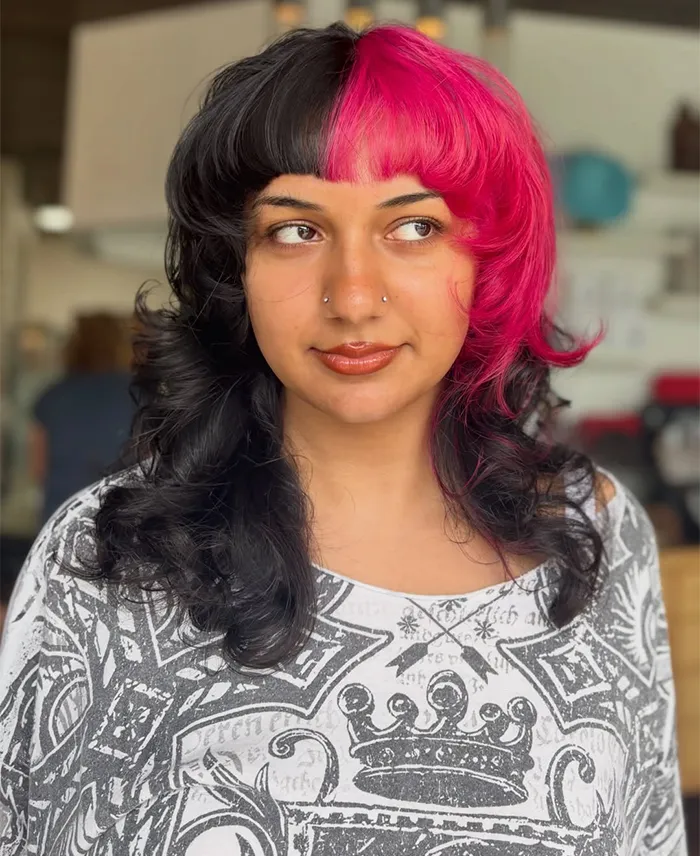 A person with a modern precision haircut and style, their curly shoulder-length hair dyed half black and half vivid pink, wears a patterned white and black top and looks slightly to the side. - Garbo A Salon & Spa TX