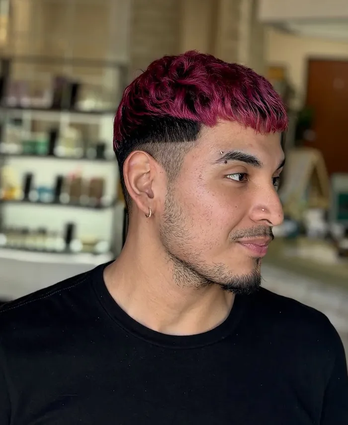 Man with short, textured magenta hair from a vivid hair color transformation, featuring a shaved line design, light beard, hoop earring, and eyebrow piercing, wearing a black shirt in an indoor setting. - Garbo A Salon & Spa TX