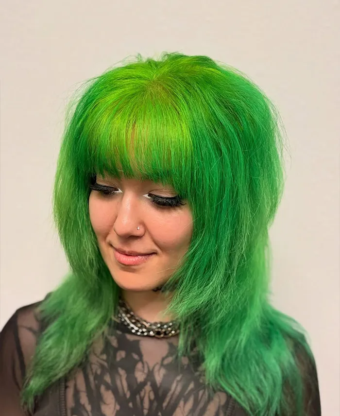 A person with layered bright green hair and bangs, styled in a modern precision haircut, looks down and smiles slightly. They are wearing a black sheer top and a chain necklace. - Garbo A Salon & Spa TX