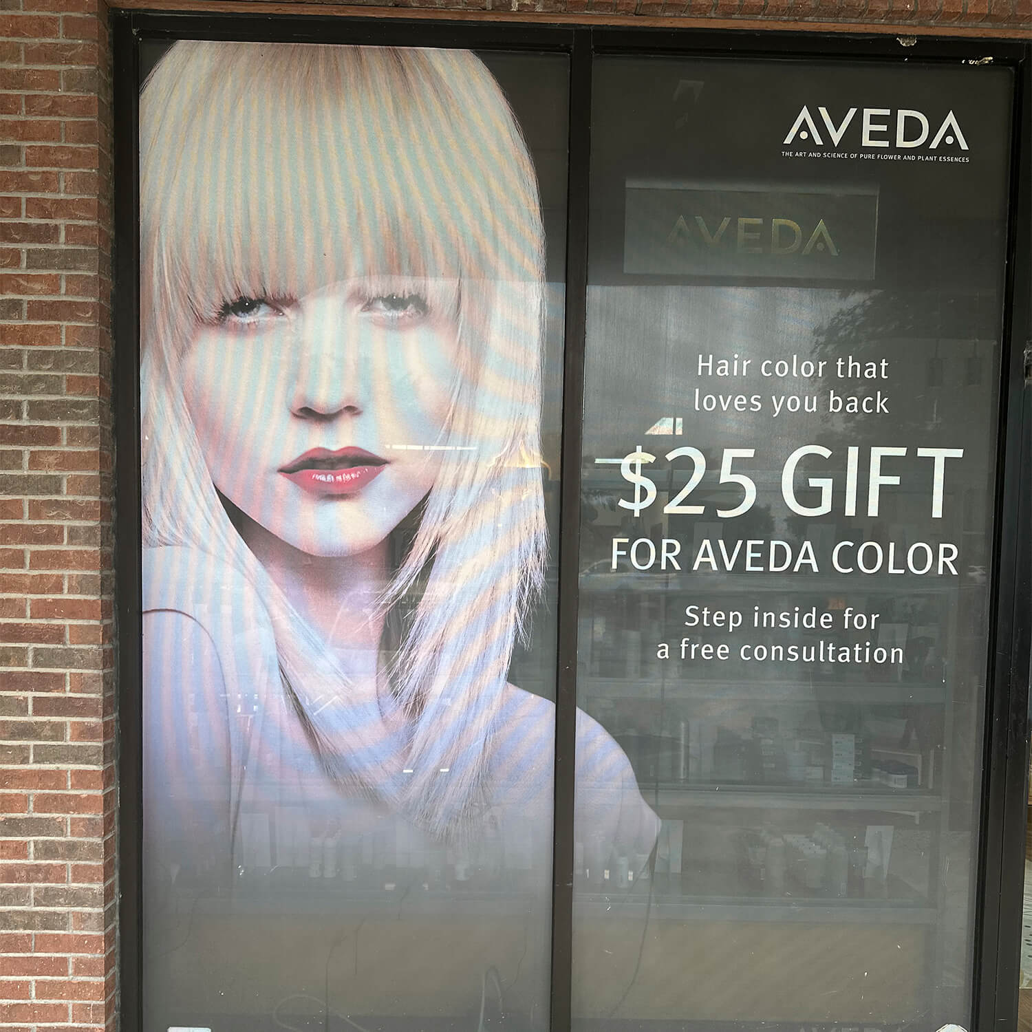 A salon window displays an Aveda advertisement featuring a woman with blonde hair and promotes a $25 gift for Aveda color with a free consultation at this eco friendly beauty salon spa. - Garbo A Salon & Spa TX