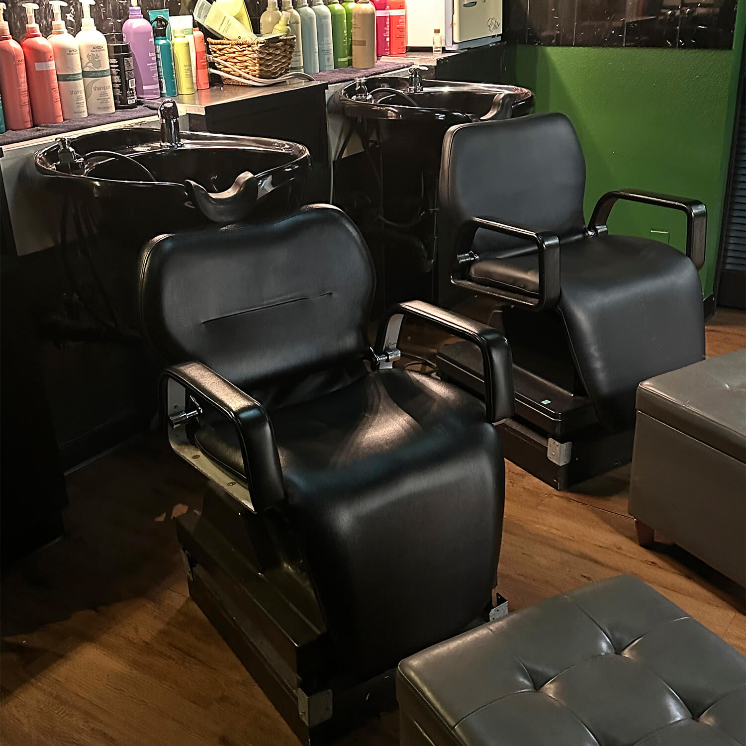 Two black salon chairs with sinks behind them, surrounded by hair care products, in a dimly lit hair salon with wooden flooring—perfect for a modern precision haircut and style. - Garbo A Salon & Spa TX