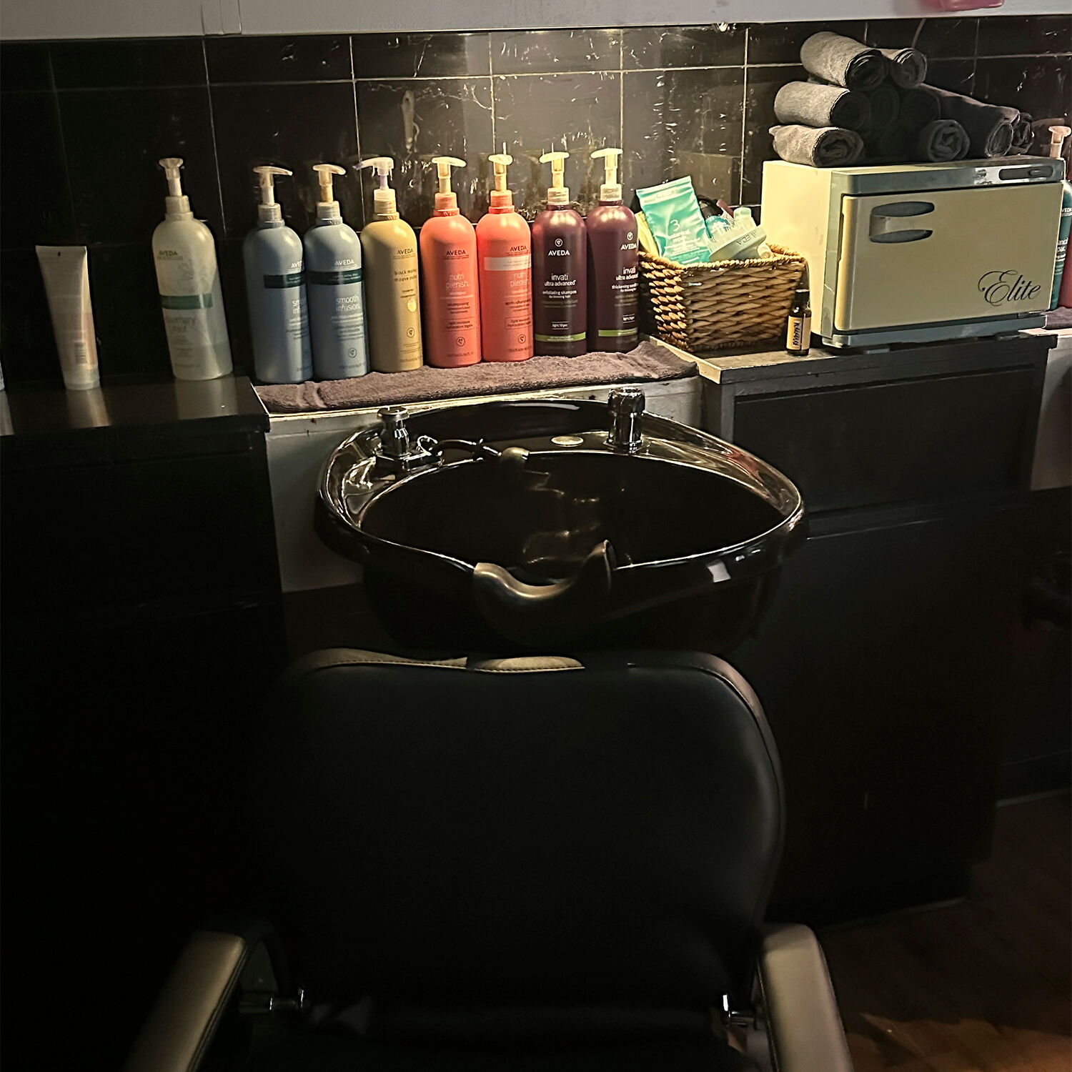 A salon station with a black chair, wash basin, assorted hair product bottles, rolled towels, and a towel warmer on a black counter—perfect for modern precision haircut and style at an eco friendly beauty salon spa. - Garbo A Salon & Spa TX