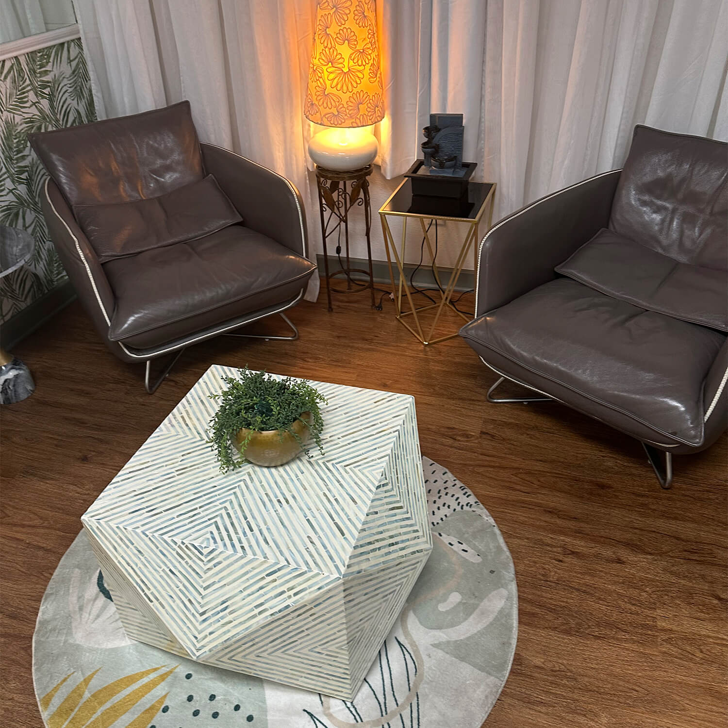 Two brown leather armchairs face a geometric white coffee table with a small plant, near a glowing lamp on a stand, all set on a round rug over wood flooring—the perfect space to discuss your next modern precision haircut and style. - Garbo A Salon & Spa TX