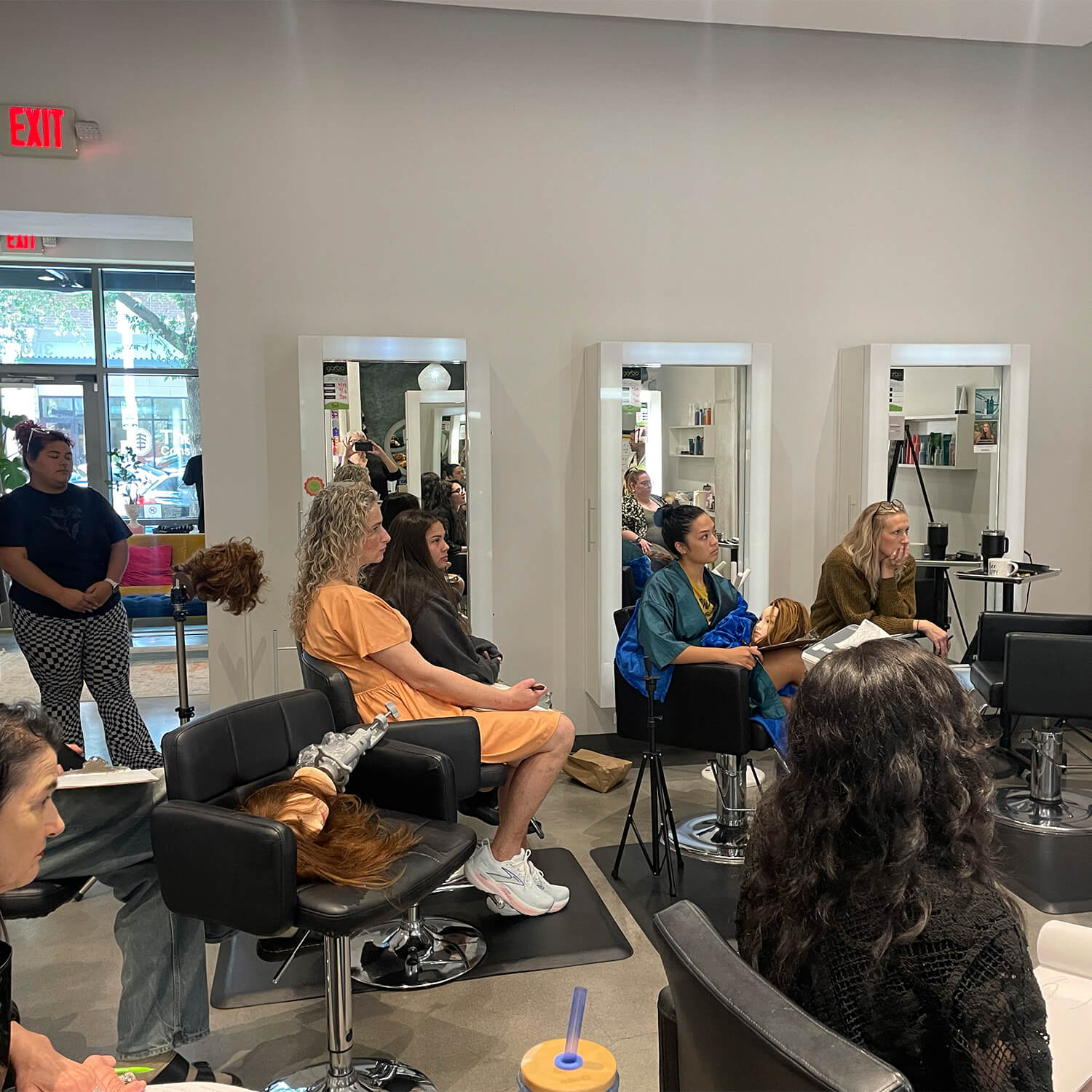 People sit in salon chairs facing forward, some with salon capes, while a few mannequin heads with wigs are placed on stands in a bright, modern Aveda salon hair styling space. - Garbo A Salon & Spa TX