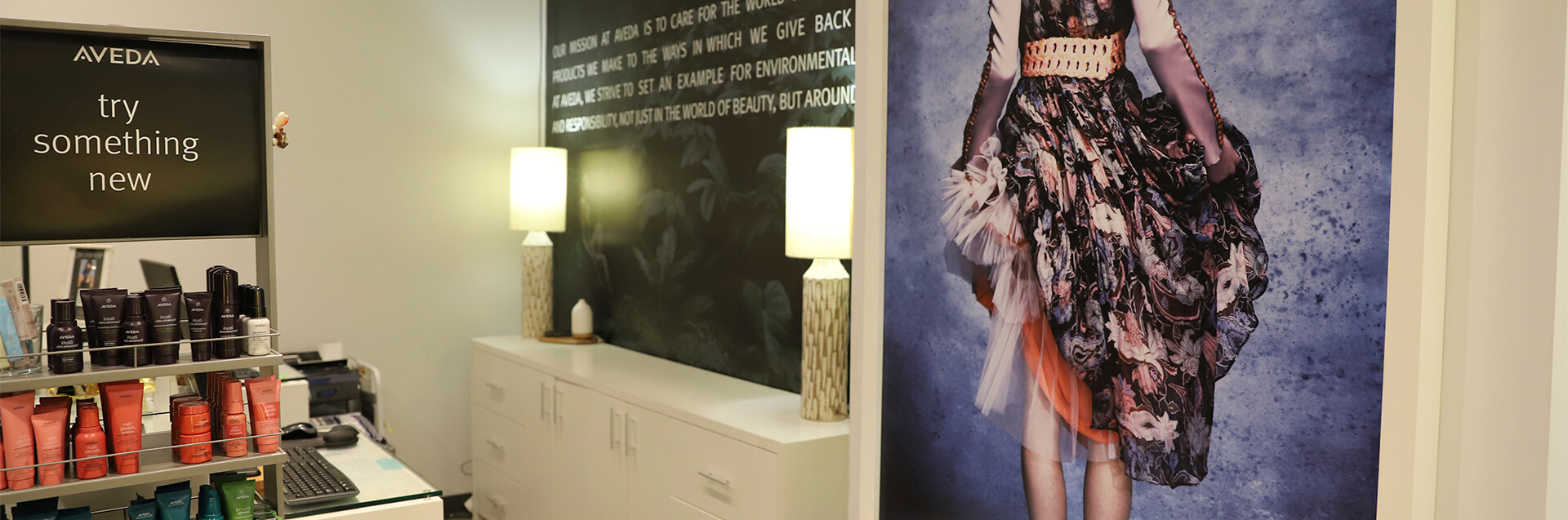 A salon interior with Aveda hair products on shelves, a wall mural of a woman in a patterned dress, white cabinets, and two table lamps—an eco friendly beauty salon spa perfect for your modern precision haircut and style. - Garbo A Salon & Spa TX