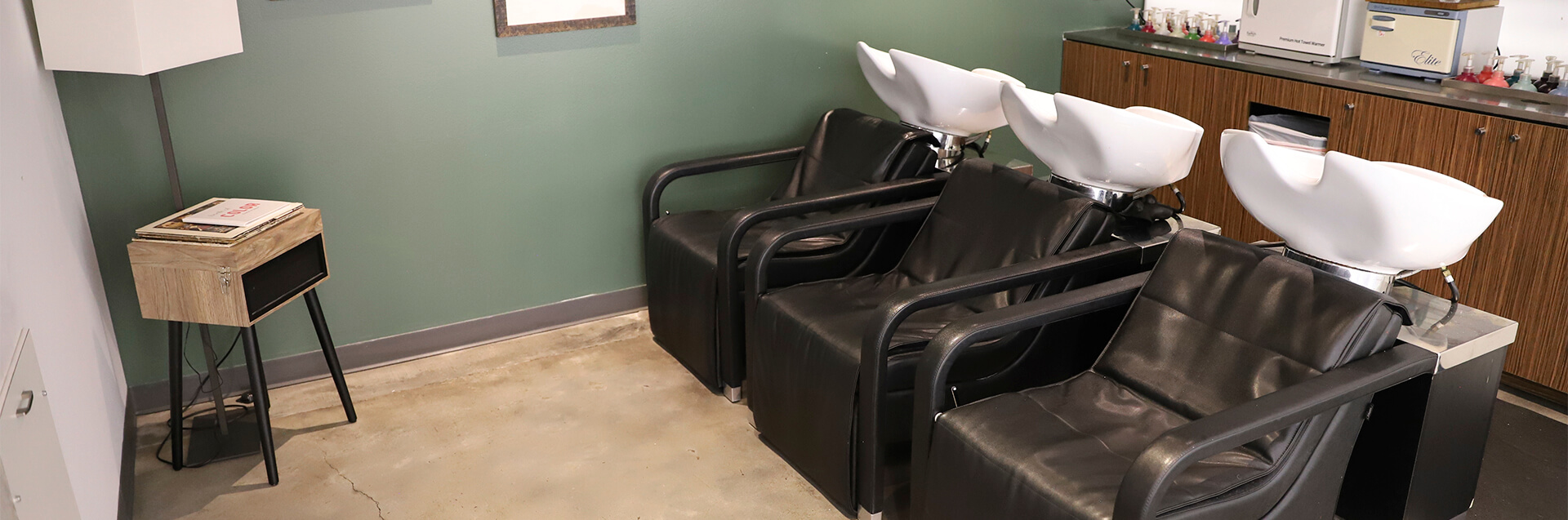 Three black salon chairs with white washbasins are lined up against a green wall in this eco friendly beauty salon spa, next to a counter with hair products and a small table with magazines. - Garbo A Salon & Spa TX