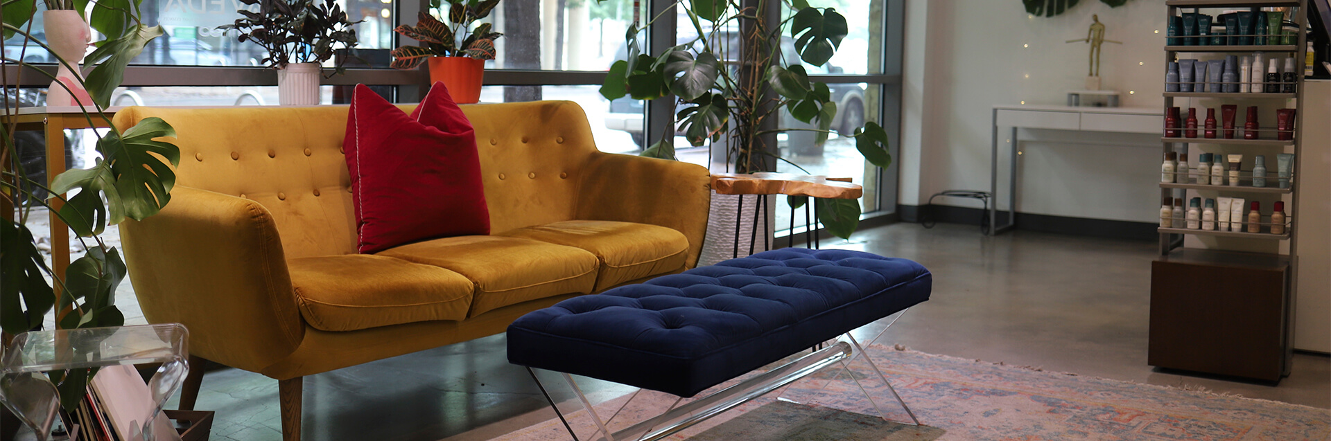 A modern, eco-friendly beauty salon spa waiting area with a yellow sofa, red pillow, blue bench, lush plants, a rug, and shelves of beauty products—perfect for your vivid hair color transformation near large windows. - Garbo A Salon & Spa TX