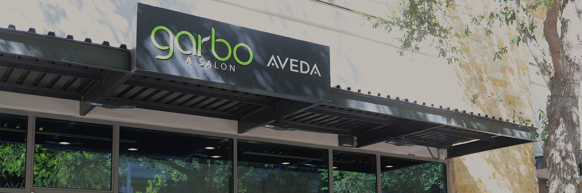 Exterior of Garbo A Salon Aveda, a top salon for hair highlights and modern precision haircut and style, with a sleek black sign above large windows, surrounded by trees and a stone wall. - Garbo A Salon & Spa TX