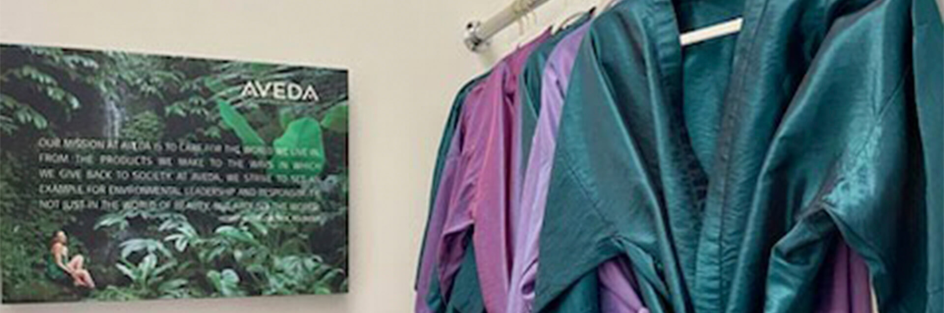 A rack of colorful robes hangs next to a wall sign with the Aveda logo and mission statement, set against lush green foliage—reflecting this eco friendly beauty salon spa’s commitment to natural care and vibrant style. - Garbo A Salon & Spa TX