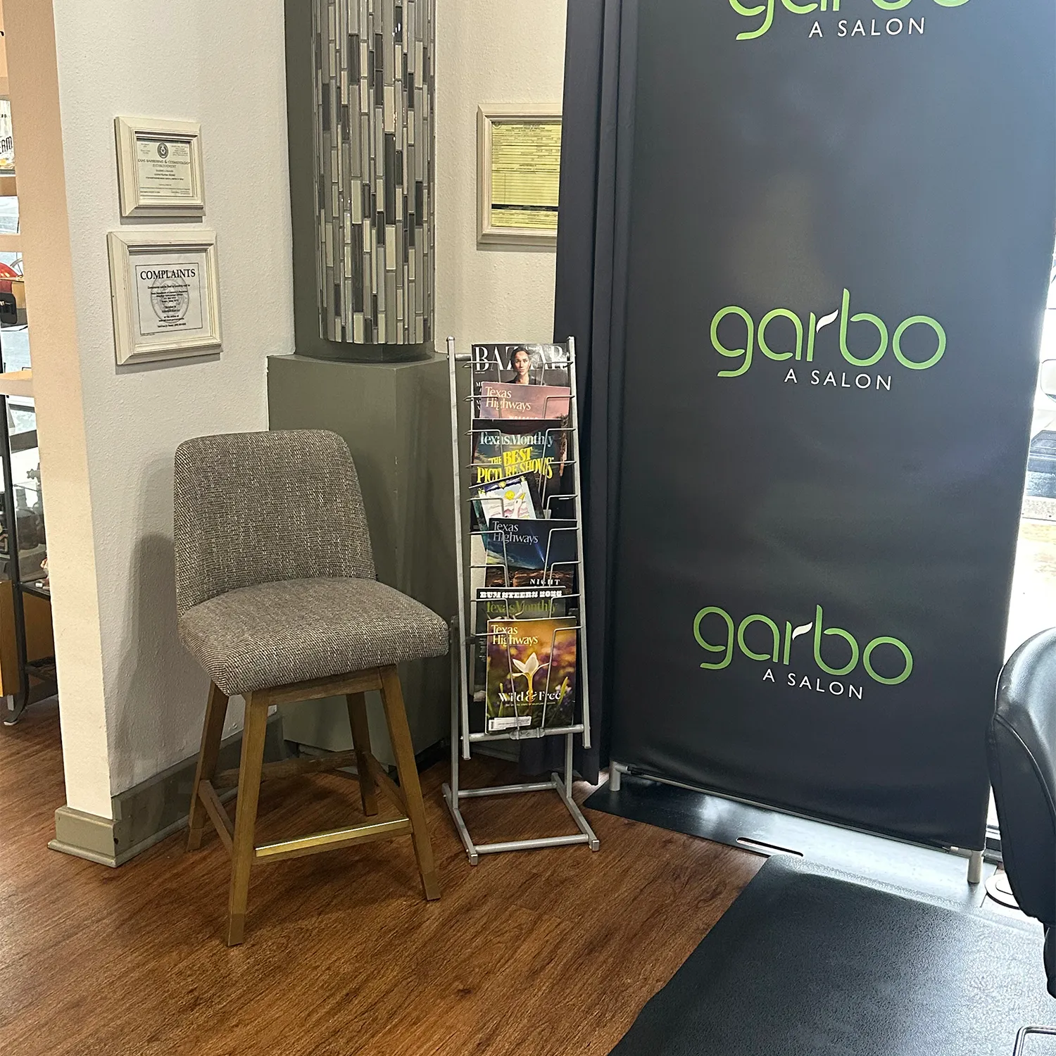 A waiting area in a salon with a gray chair, a rack of magazines, framed certificates on the wall, and a black "garbo A SALON" banner. - Garbo A Salon & Spa TX
