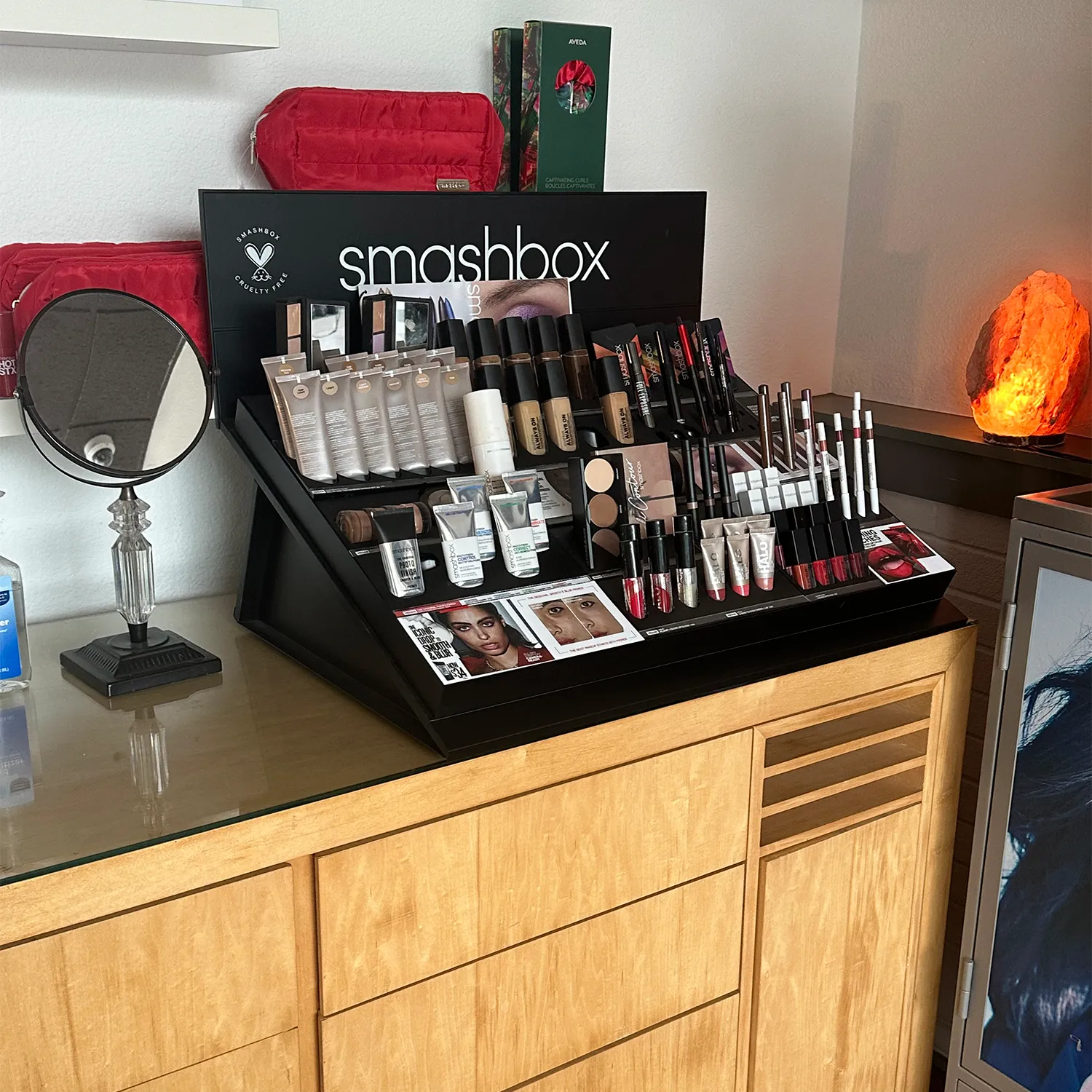 A Smashbox makeup display on a wooden cabinet with various foundations, primers, lipsticks, and eyeliners, next to a round mirror and a Himalayan salt lamp. - Garbo A Salon & Spa TX