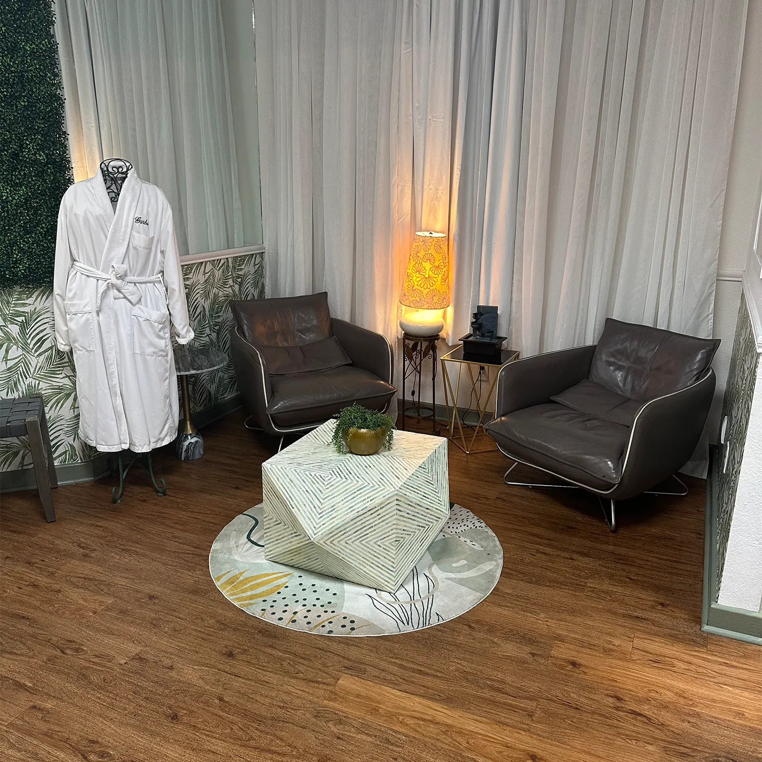 A modern waiting area with two gray armchairs, a geometric coffee table, a mannequin wearing a white robe, and a decorative lamp, all set on a wood floor with patterned walls and curtains. - Garbo A Salon & Spa TX