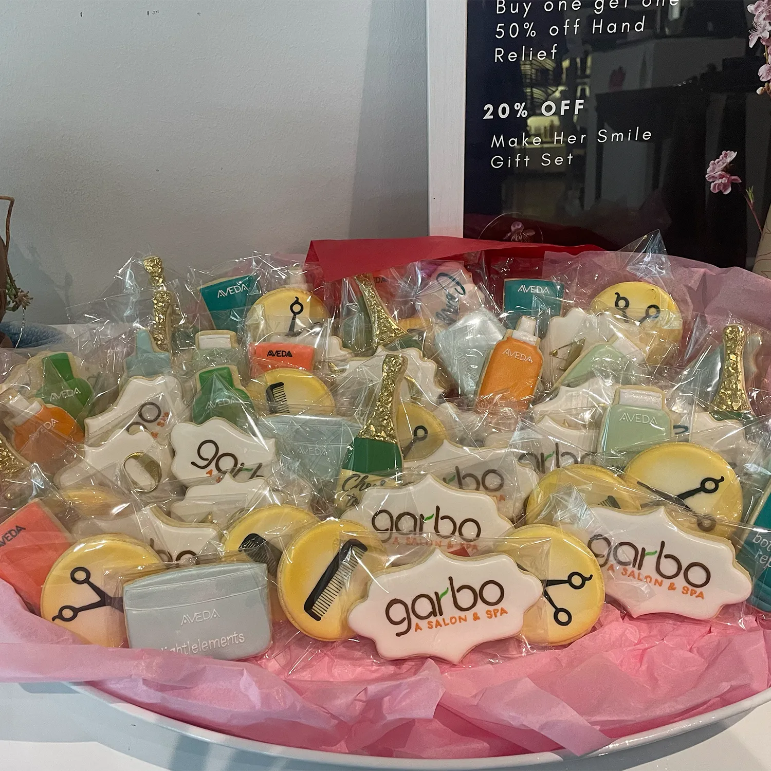 A basket of decorated cookies featuring Garbo Salon & Spa logos, hairdresser tools, Aveda products, and champagne bottles, arranged on pink tissue paper. - Garbo A Salon & Spa TX