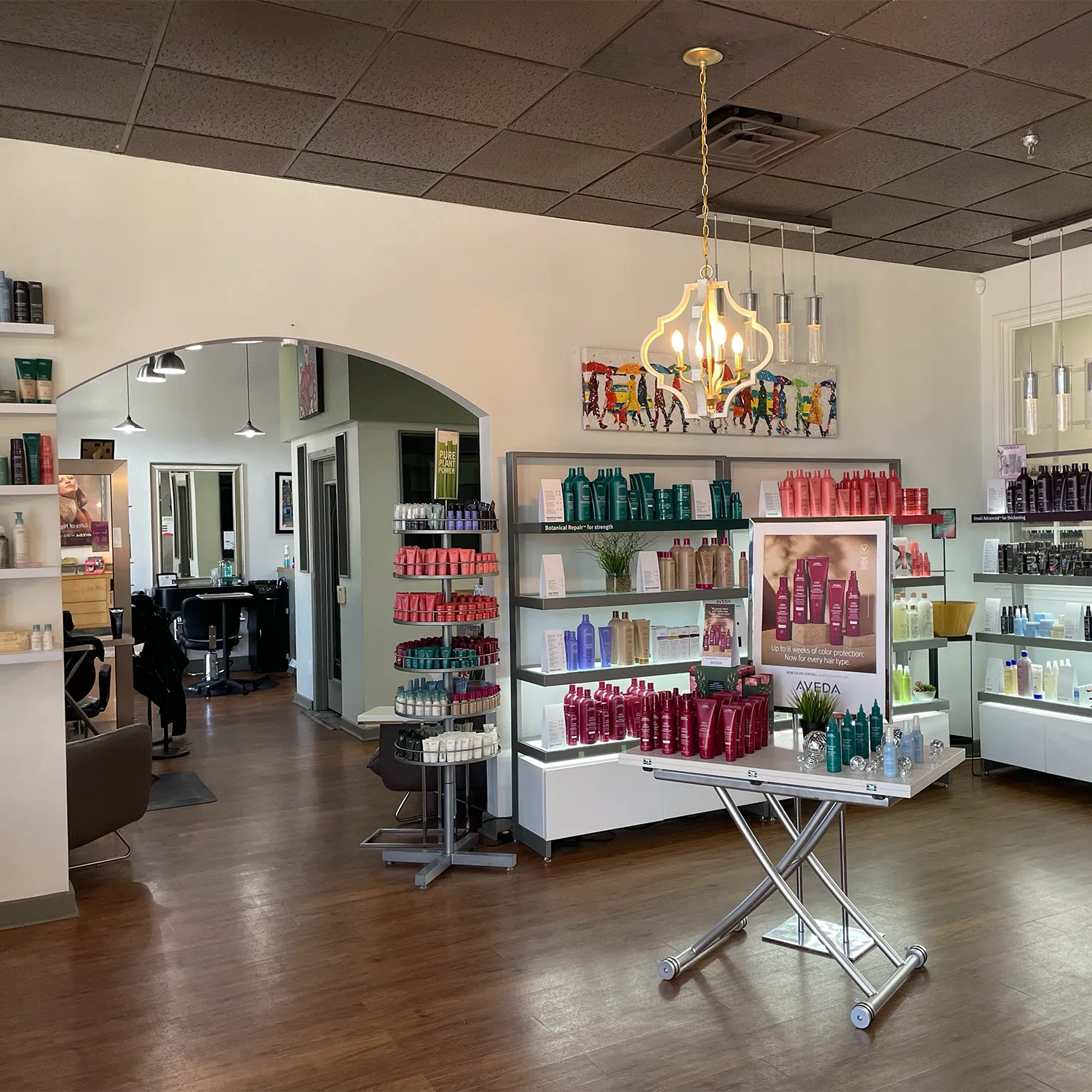 A hair salon interior with shelves displaying various hair care products, a modern chandelier, wooden floors, and salon chairs visible in the background. - Garbo A Salon & Spa TX
