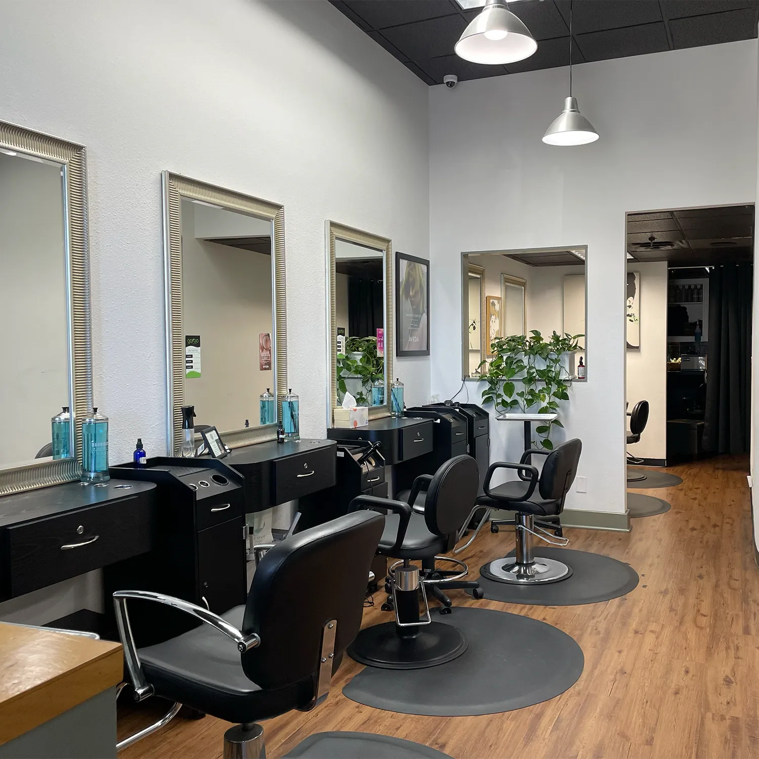 A modern hair salon with black styling chairs, mirrors, and stations along one wall, potted plants, and wooden flooring under bright overhead lights. - Garbo A Salon & Spa TX