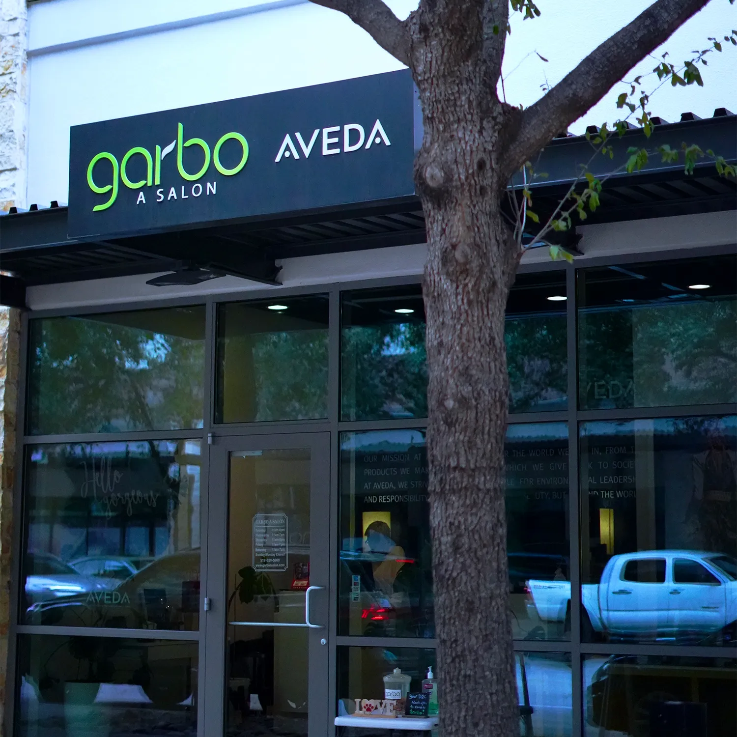 The exterior of Garbo A Salon Aveda, showing the storefront with a tree in front and reflections of cars in the windows. - Garbo A Salon & Spa TX