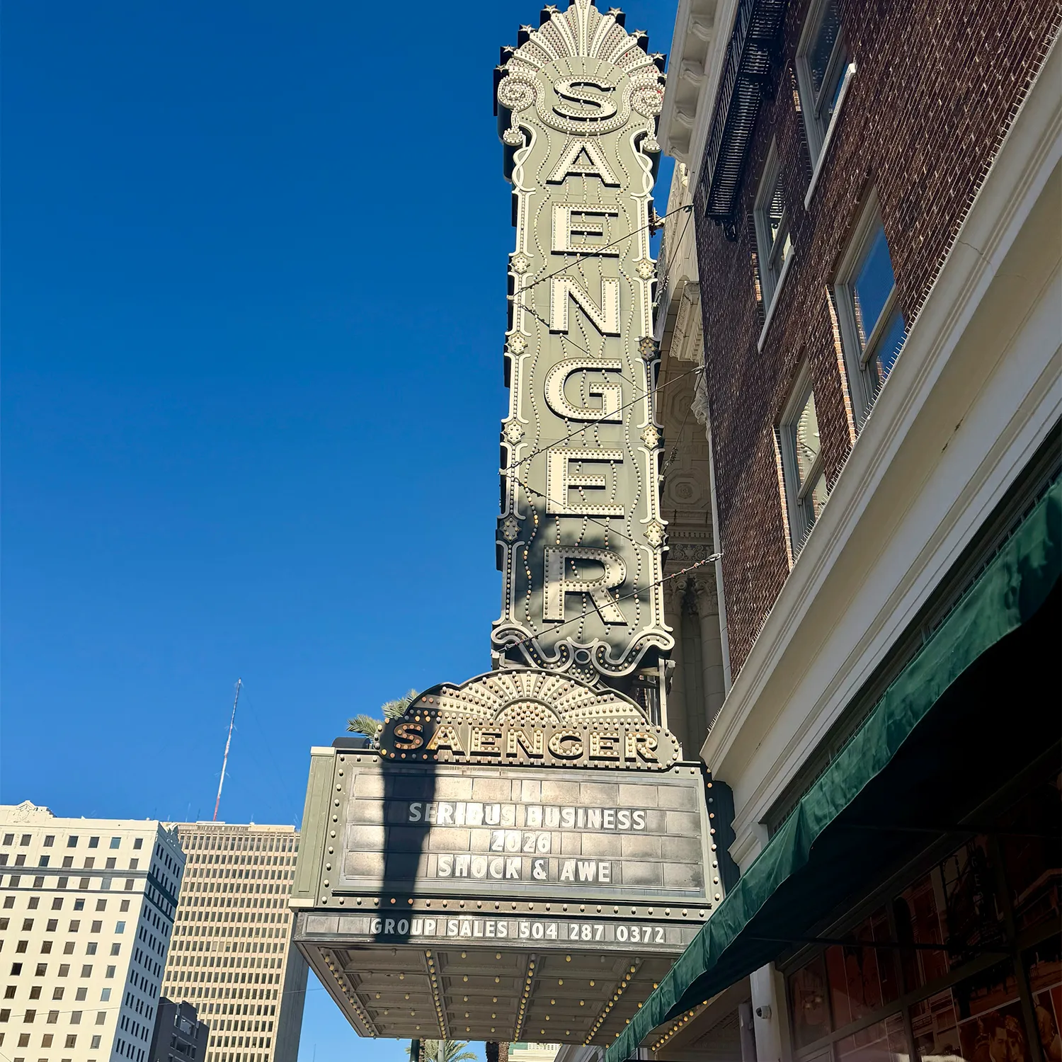 Vertical Saenger Theater sign and marquee in daylight, with the message “Show Business is in Shock & Awe” displayed above contact information. - Garbo A Salon & Spa TX