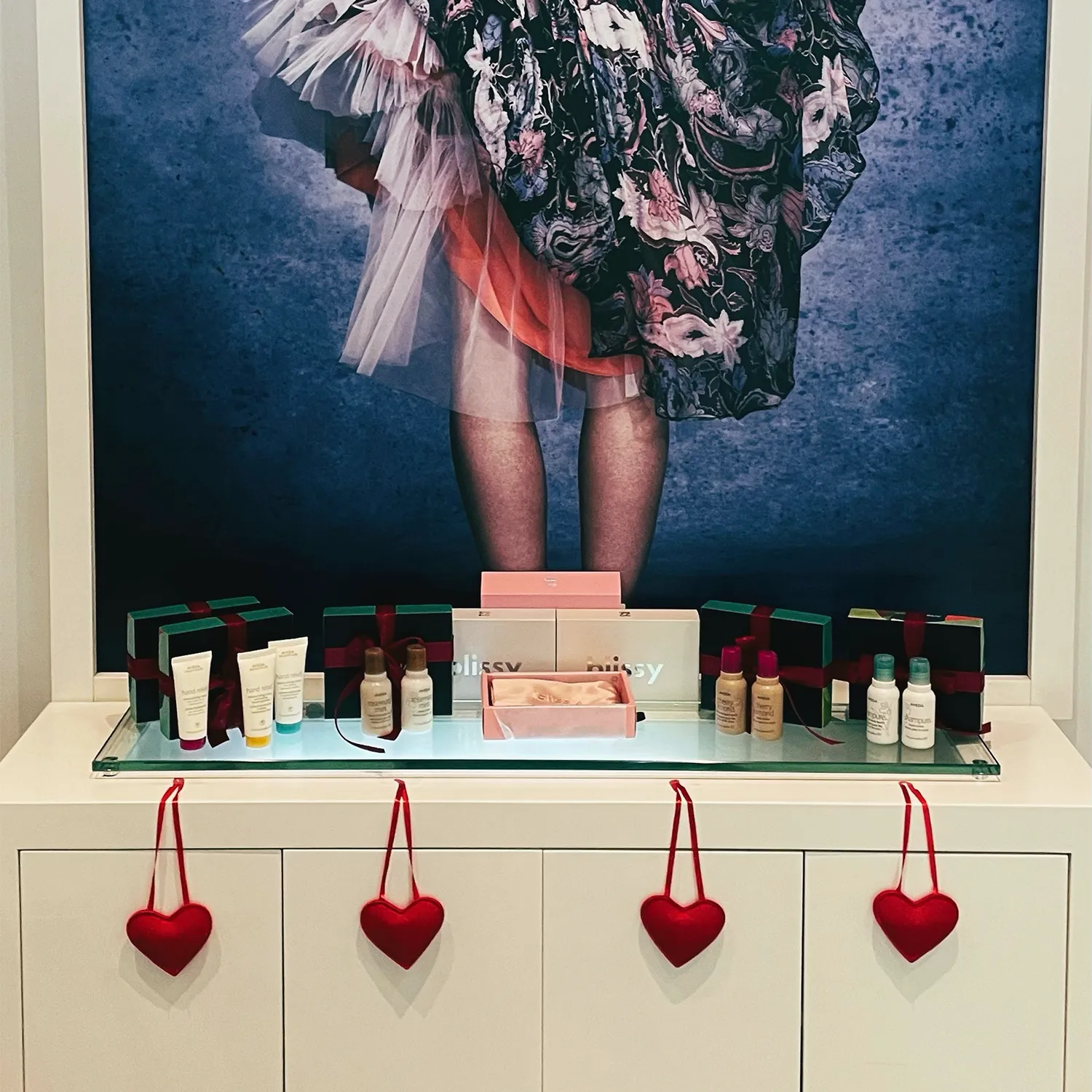 A display of skincare and beauty products beneath a photo of a model in a floral dress, with three red heart ornaments hanging from the counter. - Garbo A Salon & Spa TX
