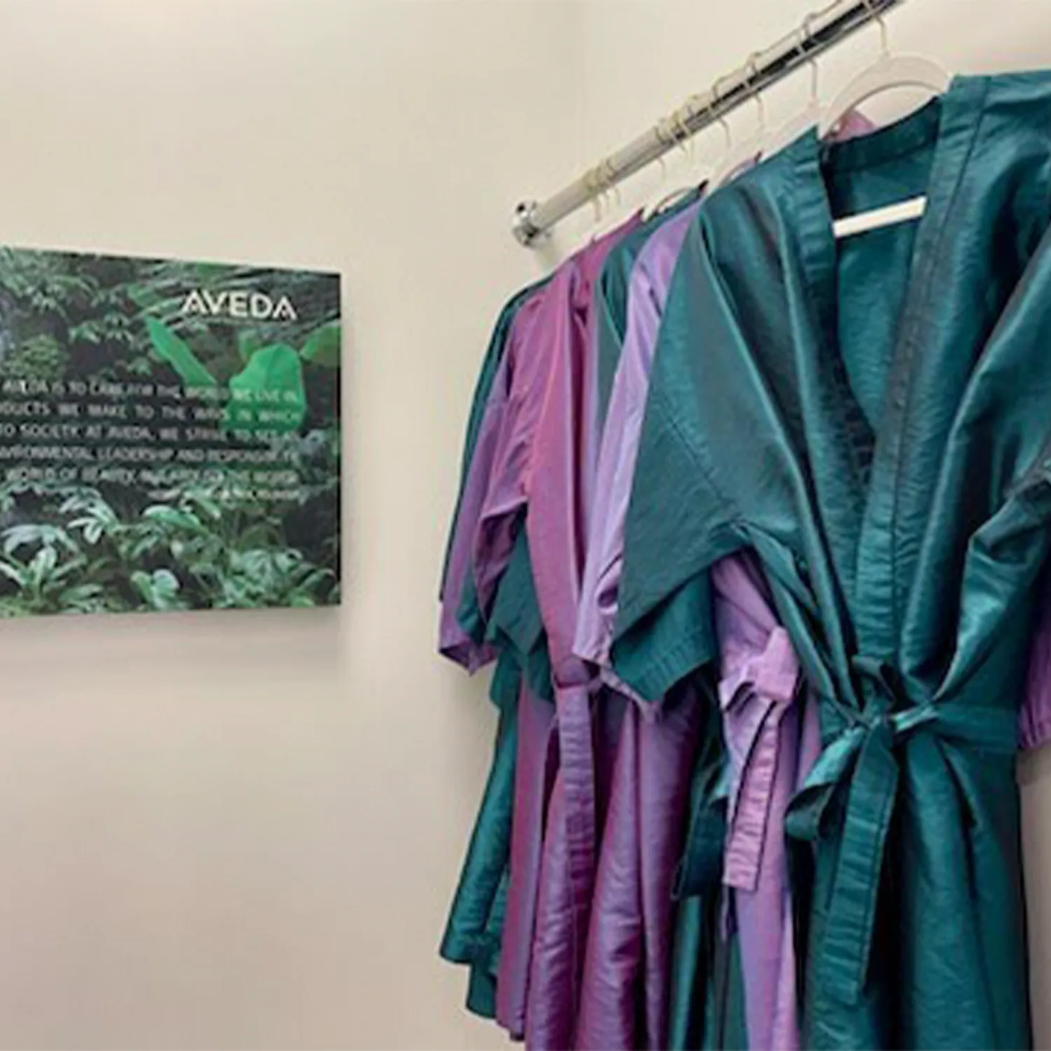 Five robes in shades of green and purple hang on a rack next to a wall sign with the Aveda logo and text, in a clean, well-lit room. - Garbo A Salon & Spa TX