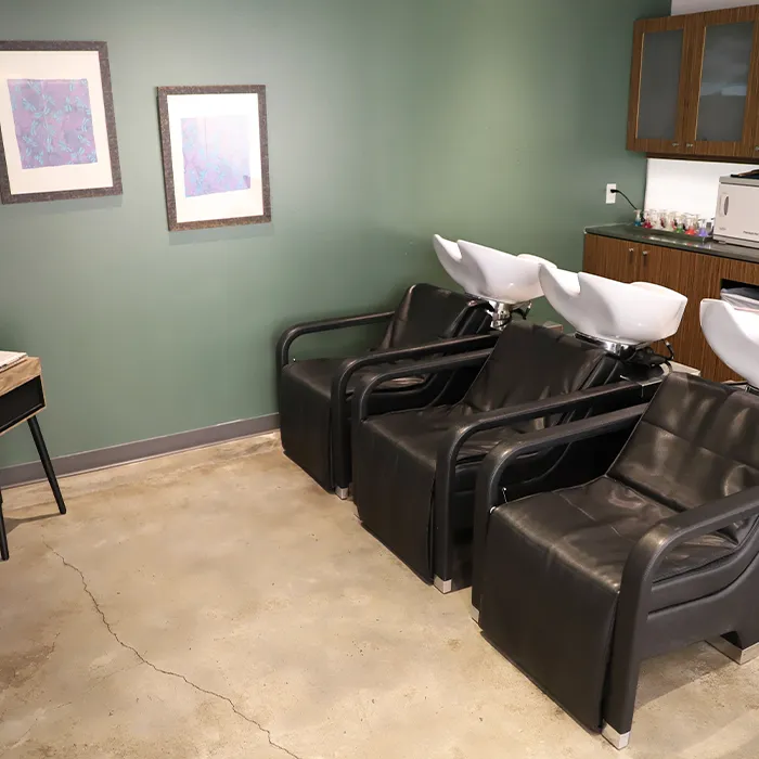 Three black salon shampoo chairs with attached white sinks are lined up against a green wall in a modern salon, with art on the wall and cabinetry in the background. - Garbo A Salon & Spa TX