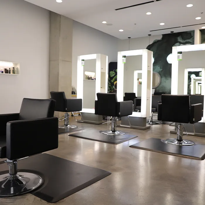 A modern hair salon with four black chairs, large illuminated mirrors, and shelves with hair products on a polished concrete floor. - Garbo A Salon & Spa TX