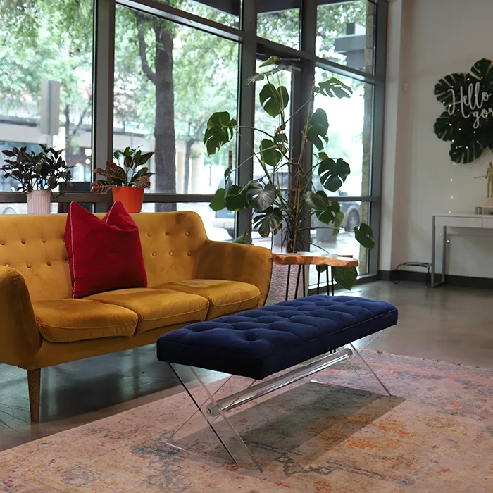 A modern living room with a yellow sofa, a red pillow, a blue bench, potted plants, and large windows showing trees outside. - Garbo A Salon & Spa TX