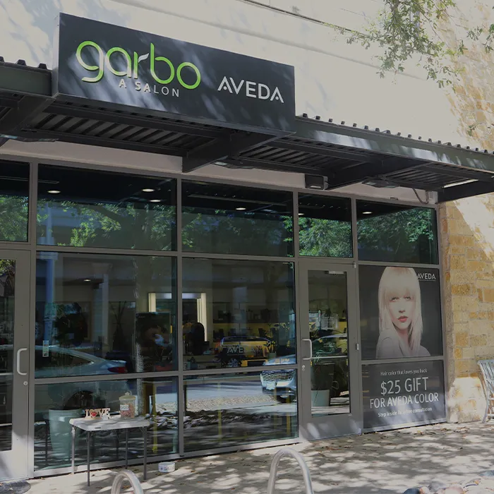 Storefront of Garbo A Salon Aveda with large windows, a black awning, and a poster advertising a $25 gift for Aveda color services. - Garbo A Salon & Spa TX