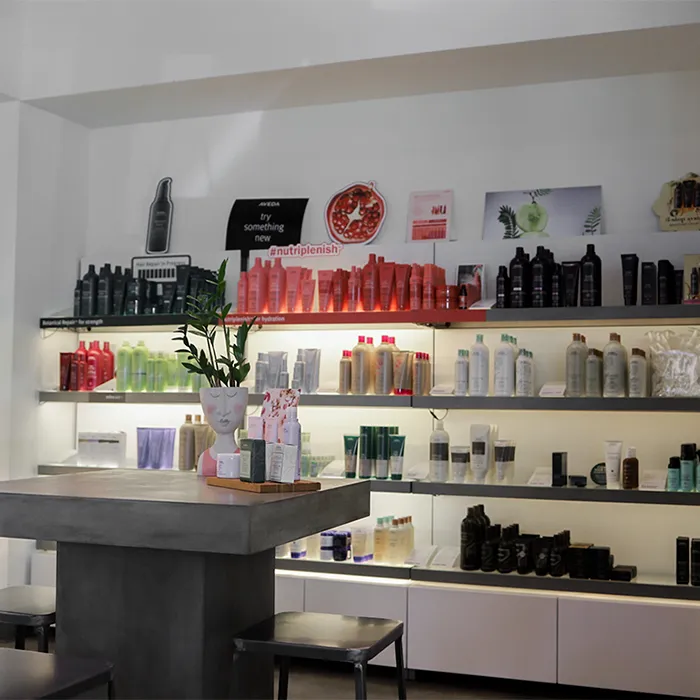 Modern salon retail area with shelves displaying various hair care products in colorful bottles, a concrete table with a plant centerpiece, and minimalist decor. - Garbo A Salon & Spa TX
