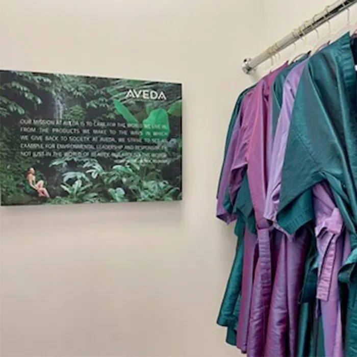 A row of colorful scrubs hangs on a rack next to a wall sign with the Aveda logo and a mission statement against a green forest background. - Garbo A Salon & Spa TX
